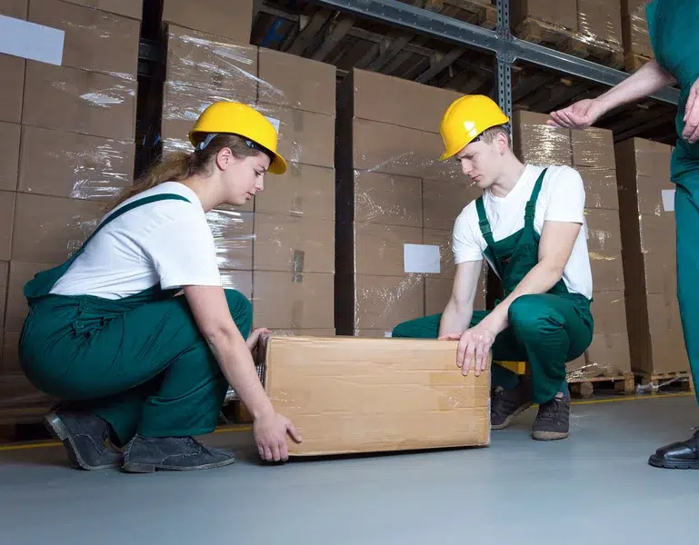 Zwei Lagerarbeiter heben gemeinsam einen Karton im ergonomischen Hockgriff – Symbolbild für muskuloskelettale Belastungen am Arbeitsplatz